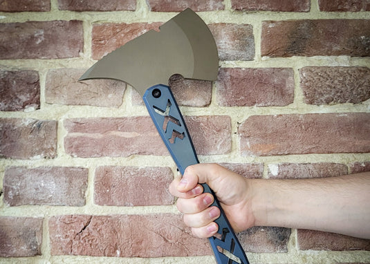 Hand holding a tactical axe with a blue handle against a brick wall, featuring the brand 'CALT'.